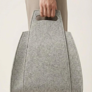 May include: A light gray felt tote bag with a structured, trapezoidal silhouette. The bag features integrated cut-out handles at the top and visible vertical seams along the front panels, creating a clean, architectural shape with a textured finish.