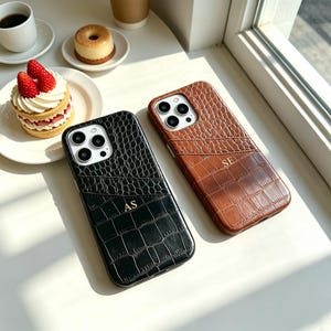 Peut inclure: Deux coques de smartphone rectangulaires avec un motif texturé en peau de crocodile. L'une est noire avec les initiales dorées AS, et l'autre est marron avec les initiales dorées SE. Les deux présentent une fente pour carte et des découpes.