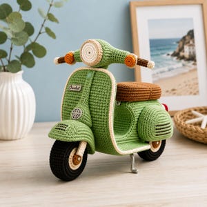Puede incluir: Un scooter de ganchillo hecho a mano en color verde claro con asiento marrón, neumáticos negros y detalles en color crema. El frente presenta un faro circular, intermitentes naranjas y una pequeña etiqueta rectangular con la palabra Crochet.