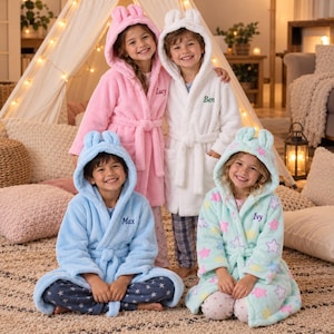Pode incluir: Quatro roupões de pelúcia com capuz e orelhas de animal. Um é rosa com o nome Lucy, um é branco com o nome Ben, um é azul claro com o nome Max e um é verde menta com um padrão de estrelas coloridas e o nome Ivy.