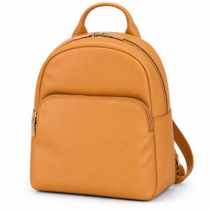 Puede incluir: Una mochila de color amarillo mostaza con un acabado liso y mate. La bolsa cuenta con un asa superior redondeada, un compartimento principal con cremallera y un bolsillo frontal más pequeño con cremallera. Herrajes metálicos dorados.