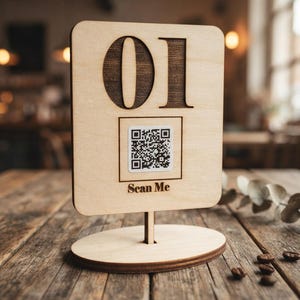 Puede incluir: Un letrero de mesa de madera clara con esquinas redondeadas, que presenta el número 01 grabado en oscuro en la parte superior. Debajo del número hay una pegatina de código QR cuadrada y el texto Scan Me grabado en madera oscura, sobre una base redonda.