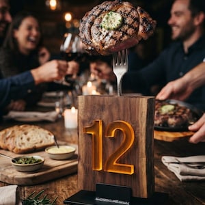 May include: A rectangular wooden table number sign featuring the number 12 in translucent orange acrylic, mounted on a black metal base. A silver fork is inserted into the top of the wood, holding a grilled steak topped with herb butter.