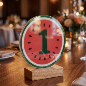 May include: A circular table number sign featuring a watermelon graphic with a red center, black seeds, and a green rind border. The number 1 is printed in dark green in the center. The sign is mounted on a rectangular light wood base.