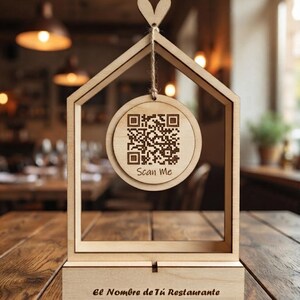 Puede incluir: Un marco de madera clara con forma de casa y una base rectangular que presenta el texto "El Nombre de Tú Restaurante". Un disco circular de madera cuelga del centro superior mediante un cordel, mostrando un código QR y el texto "Scan Me".