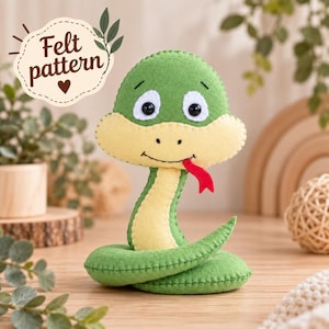 May include: A green and yellow felt snake toy with black eyes, a stitched mouth, and a red tongue. A white scalloped label in the upper left corner features the text Felt pattern with a small heart and leaf illustrations.