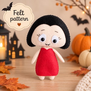 May include: A handcrafted felt doll featuring a pale face, black hair, and a red dress. The doll has large white eyes with black pupils, small eyebrows, and two tiny fangs. A decorative sign above the doll displays the text Felt pattern.