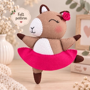 May include: A brown and white felt animal doll with a pink tutu and a small pink flower on its head. The doll has stitched facial features, including closed eyes and a nose. A label in the upper left corner reads Felt pattern.