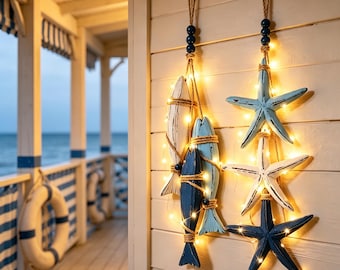 May include: Two vertical wall hangings featuring nautical motifs. One strand displays three wooden fish in white, light blue, and dark blue, while the other features three starfish in light blue, white, and dark blue, all illuminated by warm string lights.