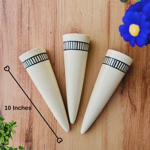 Könnte beinhalten: Drei cremefarbene, kegelförmige Wandvasen mit einem schwarzen vertikalen Streifenmuster am Rand. Jede Vase ist 25,4 cm lang, wie durch die grafische Linie angezeigt. Die Vasen haben ein glattes, konisches Design und eine weite Öffnung.
