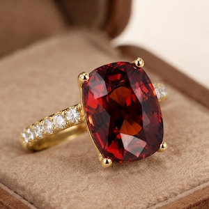 Puede incluir: Un anillo de oro con una gran piedra preciosa de color rojo intenso en talla cojín, sujeta por cuatro garras. La banda está hecha de oro amarillo y decorada con una hilera de pequeñas piedras blancas redondas y transparentes.