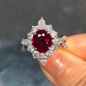 Puede incluir: Un anillo de tono plateado con un gran rubí rojo oscuro de corte ovalado en el centro. El rubí está rodeado por un halo de pequeños diamantes blancos brillantes en cortes redondos y marquesa. La banda tiene un diseño trenzado con diamantes.