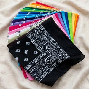 May include: A collection of square bandanas arranged in a fan shape, featuring a black bandana with a white paisley pattern on top. The stack includes various solid colors such as red, orange, yellow, green, blue, purple, pink, white, and brown.
