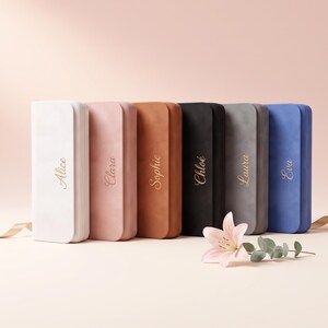 Peut inclure: Six étuis rectangulaires de couleurs blanc, rose, beige, noir, gris et bleu, chacun portant un prénom doré : Alice, Clara, Sophie, Chloé, Laura et Eva. Un lys rose pâle avec des feuilles vertes est posé devant les étuis.