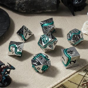 May include: A set of seven polyhedral dice made of polished silver-toned metal. Each die features a recessed, winding green serpent design snaking between the black engraved numbers. The set includes a d4, d6, d8, d10, d12, d20, and a percentile die.