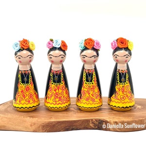 May include: Four wooden dolls with black hair and painted faces, wearing yellow and red patterned dresses, black necklaces, and colorful flower crowns. The dolls are standing on a wooden base.