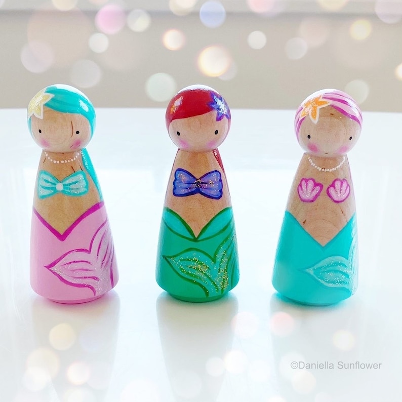 Puede incluir: Tres mu&ntilde;ecas de madera con forma de clavija vestidas de sirenas. Las mu&ntilde;ecas tienen caras y cabello pintados y llevan colas de sirena con detalles de purpurina. Una mu&ntilde;eca tiene detalles rosa y azul, otra tiene detalles verde y azul y la &uacute;ltima tiene detalles azul turquesa y rosa.