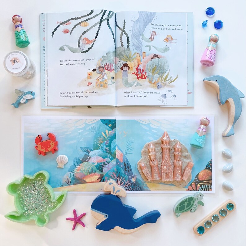 Puede incluir: Un conjunto de juguetes con tem&aacute;tica oce&aacute;nica para ni&ntilde;os, que incluye una ballena azul, una tortuga verde, un cangrejo rojo, un castillo de arena rosa y un delf&iacute;n azul. Los juguetes est&aacute;n dispuestos sobre una superficie blanca con un libro sobre el oc&eacute;ano en el fondo.