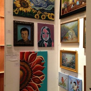 May include: A gallery wall display featuring multiple framed and unframed paintings, including a large sunflower and starry night scene, a portrait of a person, a stylized face, a close-up of a sunflower, a park scene, a dog, and bird paintings.