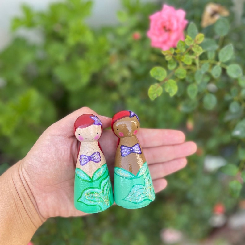 Puede incluir: Dos mu&ntilde;ecas de madera con forma de pe&oacute;n vestidas de sirenas. Una mu&ntilde;eca tiene el pelo rojo y una cola verde con purpurina dorada. La otra mu&ntilde;eca tiene la piel morena y una cola verde con purpurina dorada. Ambas mu&ntilde;ecas tienen lazos morados.