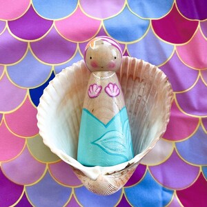 Puede incluir: Una mu&ntilde;eca de madera pintada para parecer una sirena, con un vestido turquesa y blanco, con conchas rosadas en el pecho. La mu&ntilde;eca est&aacute; sentada dentro de una concha, sobre un fondo rosa y morado.