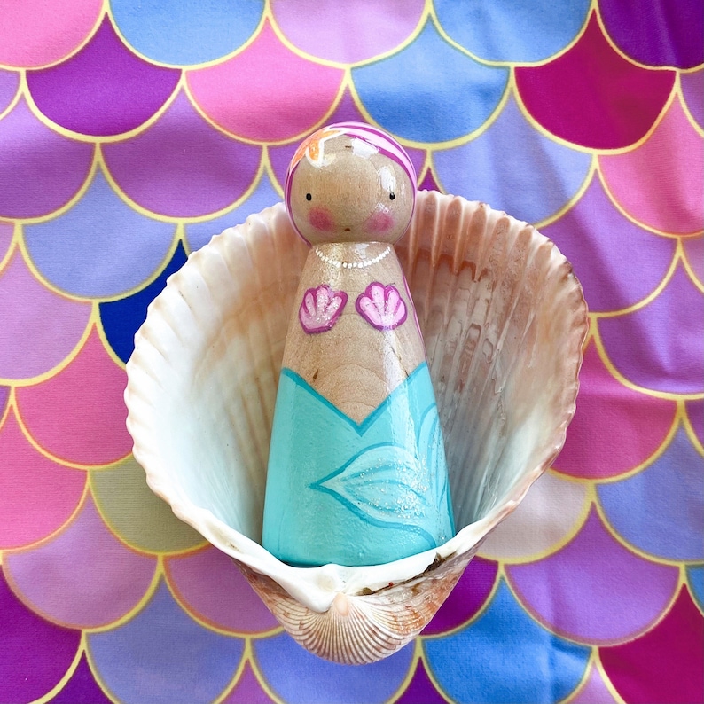 Puede incluir: Una mu&ntilde;eca de madera pintada para parecer una sirena, con un vestido turquesa y blanco, con conchas rosadas en el pecho. La mu&ntilde;eca est&aacute; sentada dentro de una concha, sobre un fondo rosa y morado.