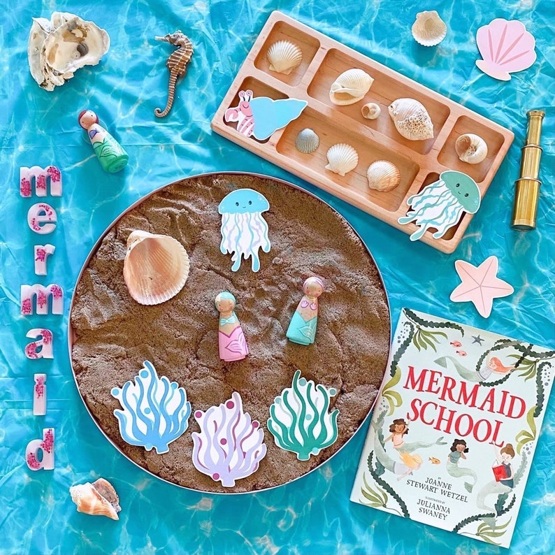Puede incluir: Una bandeja marr&oacute;n llena de arena, rodeada de varios juguetes con tem&aacute;tica de sirenas, incluyendo un libro titulado "Mermaid School", mu&ntilde;ecas de madera, conchas y medusas.