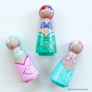 Puede incluir: Tres mu&ntilde;ecas de madera con forma de clavija vestidas de sirenas. Las mu&ntilde;ecas est&aacute;n pintadas en tonos de rosa, verde y azul. Cada mu&ntilde;eca tiene un peinado y rasgos faciales diferentes. Las mu&ntilde;ecas son una adici&oacute;n divertida y caprichosa a la sala de juegos de cualquier ni&ntilde;o.