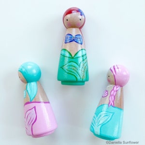 Puede incluir: Tres mu&ntilde;ecas de madera con forma de pe&oacute;n vestidas de sirenas. Una mu&ntilde;eca tiene el pelo rojo y una cola verde, otra tiene el pelo azul y una cola rosa, y la tercera tiene el pelo rosa y una cola azul.