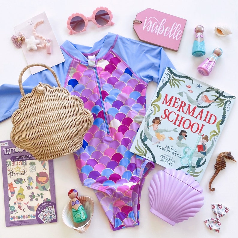Puede incluir: Un traje de ba&ntilde;o de tem&aacute;tica de sirena con un patr&oacute;n de escamas de color morado, rosa y azul. El traje de ba&ntilde;o tiene una cremallera en la parte delantera y se combina con un bolso de mimbre con forma de concha. La imagen tambi&eacute;n incluye un libro titulado "Mermaid School" y otros accesorios de tem&aacute;tica de sirena.