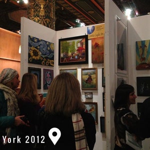 May include: An art gallery display featuring multiple framed paintings on white walls, including a sunflower piece, a street scene, and a poppy field. The text New York 2012 is visible at the bottom left with a location pin icon next to it.