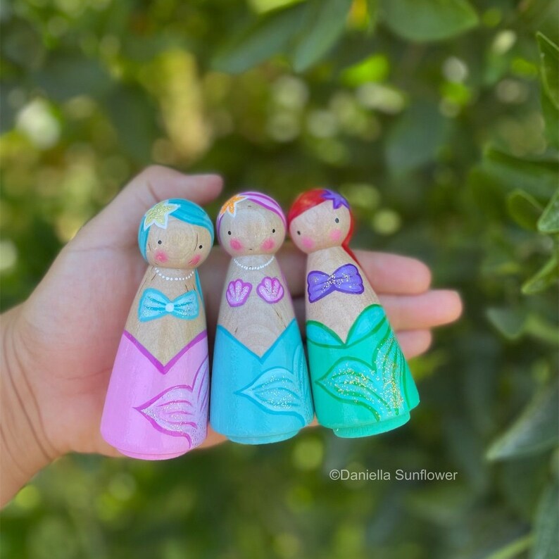 Puede incluir: Tres mu&ntilde;ecas de madera pintadas como sirenas. Las mu&ntilde;ecas llevan colas de sirena rosa, azul y verde con detalles de purpurina. Cada mu&ntilde;eca tiene un top y un pelo de color diferente.