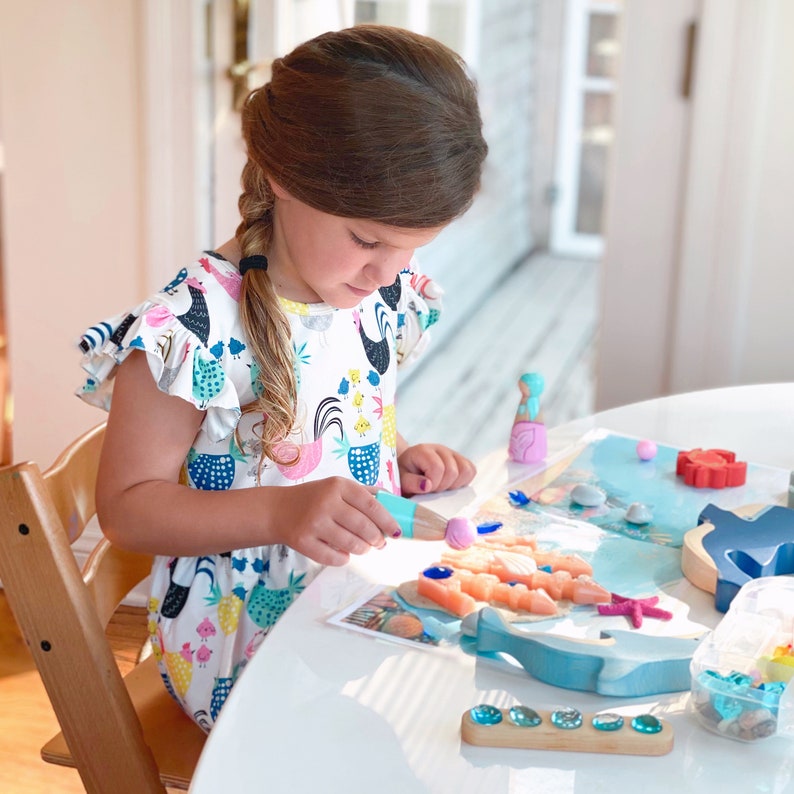 Puede incluir: Un ni&ntilde;o juega con un conjunto de juguetes de madera con tem&aacute;tica oce&aacute;nica. El conjunto incluye un delf&iacute;n, una estrella de mar, conchas marinas y una figura de sirena. El ni&ntilde;o est&aacute; usando una herramienta azul y rosa para colocar las piezas en una alfombra de juego azul y blanca.