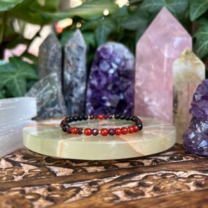 May include: A beaded bracelet featuring alternating faceted red garnet beads and smooth black onyx beads, accented with small gold-toned metal spacers. The circular jewelry piece is arranged in a repeating pattern of red and black stones.