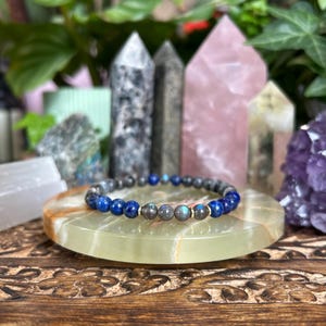 May include: A beaded bracelet featuring alternating round lapis lazuli beads in deep blue and labradorite beads with grey tones, accented by small gold-toned metal spacers. The beads are strung on an elastic cord, forming a circular shape.