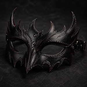 May include: A black leather mask featuring a pointed, jagged silhouette with sharp, wing-like edges and a central vertical ridge. The mask is detailed with dark red stitching along the seams and includes three small metal rivets and a side buckle strap.