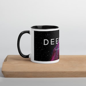 Può includere: Una tazza in ceramica bianca con manico e interno neri. L'esterno presenta una grafica di una nebulosa dello spazio profondo con nubi gassose viola e blu su uno sfondo nero stellato, con il testo bianco DEEP stampato sul lato.