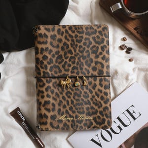 May include: A rectangular notebook with a brown and black leopard print cover featuring the name Marie Kevin embossed in gold. A black elastic band wraps around the center, holding three small gold charms: a bow, a heart, and a letter K.