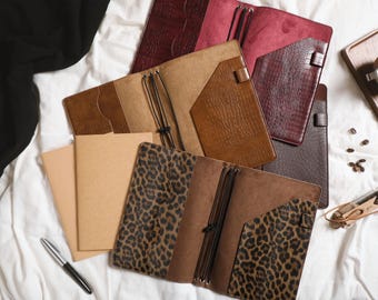 May include: A collection of open leather notebook covers in various textures and colors, including tan, dark brown, burgundy, and a leopard print pattern. Each cover features internal pockets, elastic bands for securing inserts, and a pen loop.