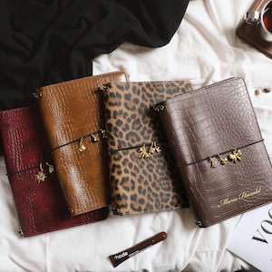 May include: Four rectangular journals with textured covers in red, tan, leopard print, and dark brown. Each journal features a black elastic band with small gold-toned charms. The dark brown journal is embossed with the name Marie Harald.