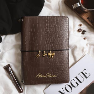 May include: A dark brown rectangular notebook with a crocodile-embossed texture. The cover features the gold-colored text AdoraHarvie and three small gold-toned charms—a letter H, a heart, and a bow—attached to a black elastic band.