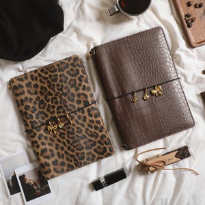May include: Two rectangular notebooks with elastic closures. One features a brown leopard print pattern, while the other has a dark brown crocodile-embossed texture. Both notebooks are adorned with small gold-toned metal charms attached to the elastic bands.