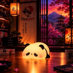 Könnte beinhalten: Eine leuchtende Panda-Lampe aus weichem Silikon mit rundem Körper, schwarzen Ohren und schwarzen Augenpartien. Die Lampe strahlt ein warmes, diffuses Licht aus und steht auf einer Holzoberfläche. Sie hat eine kleine Nase und rosafarbene Wangen.
