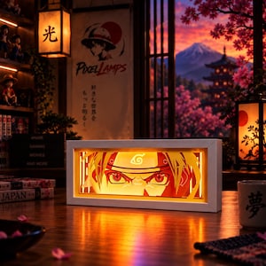 Può includere: Una scatola luminosa rettangolare con cornice bianca che presenta un design in carta tagliata a strati di un personaggio con intensi occhi rossi e una fascia con un simbolo a spirale. La calda luce arancione illumina i tratti del viso.