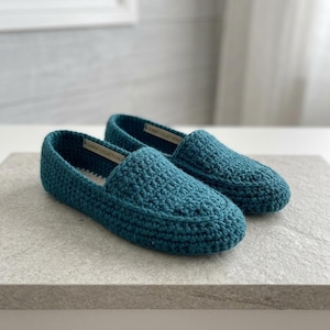 Könnte beinhalten: Ein Paar gehäkelte Slipper in Blaugrün mit einem strukturierten Strickmuster und einem kleinen weißen Innenetikett mit der Aufschrift LOKKE COLLECTION. Die Slipper haben eine abgerundete Zehenpartie und eine gleichmäßige Garnstruktur.