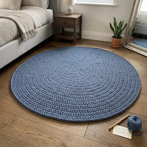 May include: A large, circular, blue crochet rug featuring a dense, textured stitch pattern. Beside the rug lies a wooden crochet hook and a small ball of matching blue yarn, along with a white card labelled Large Oval Rug Pattern.