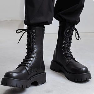Könnte beinhalten: Ein Paar schwarze, wadenhohe Kampfstiefel mit abgerundeter Zehenpartie, einer dicken, profilierten Gummisohle und einer Schnürung mit silberfarbenen Metallösen. Die Stiefel verfügen über einen vertikalen Reißverschluss an der Innenseite.