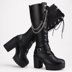 May include: Black knee-high boots with a thick platform sole and a chunky block heel. The boots feature front lace-up closures, silver-toned metal eyelets, a decorative silver chain draped across the upper shaft, and a buckled strap at the ankle.