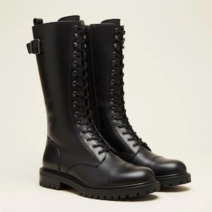 Könnte beinhalten: Ein Paar schwarze wadenhohe Stiefel mit Schnürung und Metallösen, seitlichem Reißverschluss und einem Schnallenriemen am Schaft. Die Stiefel verfügen über eine abgerundete Zehenkappe, eine Anziehschlaufe an der Ferse und eine dicke Gummiprofilsohle.