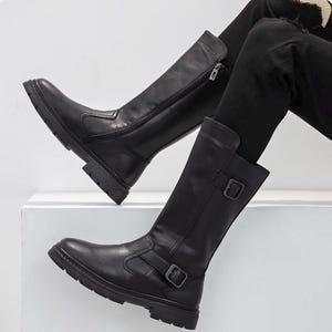 Puede incluir: Un par de botas negras a media pantorrilla con acabado liso, suela gruesa dentada y detalle de correa con hebilla lateral. Las botas tienen una caña estructurada con cierre de cremallera lateral visible, punta redondeada y un tacón robusto.